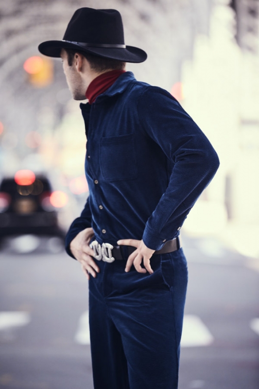 A man in a blue velvet jumpsuit and black hat stands on a city street looking away, with blurred cars and buildings in the background.