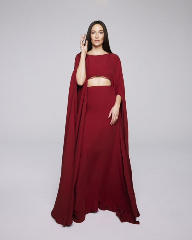 Elegant woman in a long maroon dress with cape sleeves poses in a studio with hand raised and neutral expression, full-length shot.