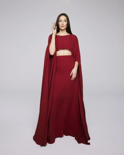 Elegant woman in a long maroon dress with cape sleeves poses in a studio with hand raised and neutral expression, full-length shot.
