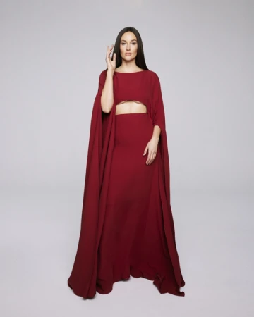 Elegant woman in a long maroon dress with cape sleeves poses in a studio with hand raised and neutral expression, full-length shot.