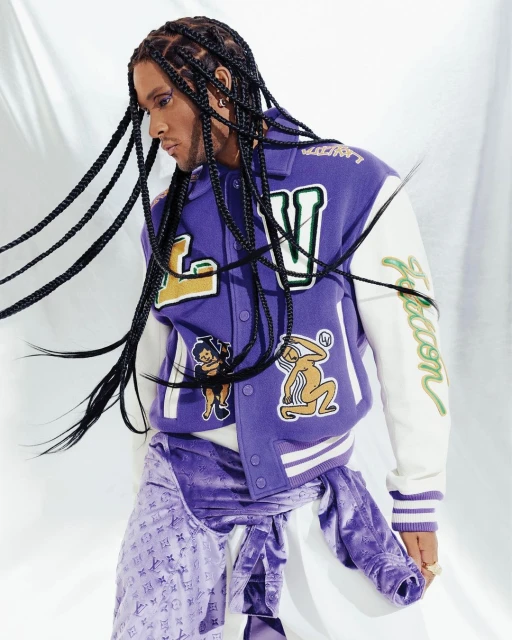 A person with braided hair, adorned in a purple varsity jacket with off-white sleeves and patterned pants against a white backdrop.