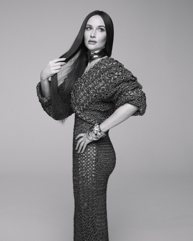 Black and white studio portrait of a brunette woman in a knitted dress and jewellery, striking a fashion pose against a neutral backdrop.