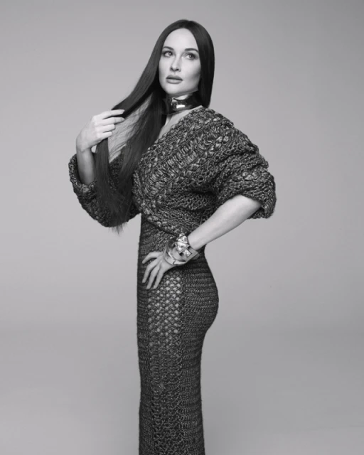 Black and white studio portrait of a brunette woman in a knitted dress and jewellery, striking a fashion pose against a neutral backdrop.