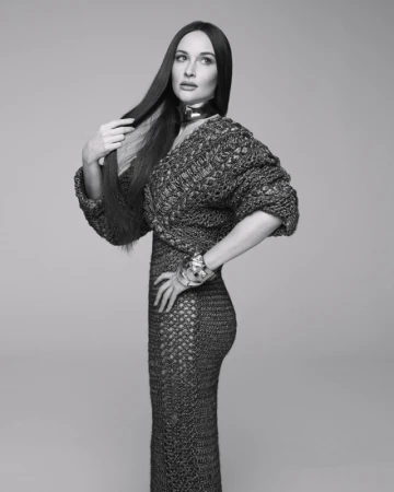 Black and white studio portrait of a brunette woman in a knitted dress and jewellery, striking a fashion pose against a neutral backdrop.