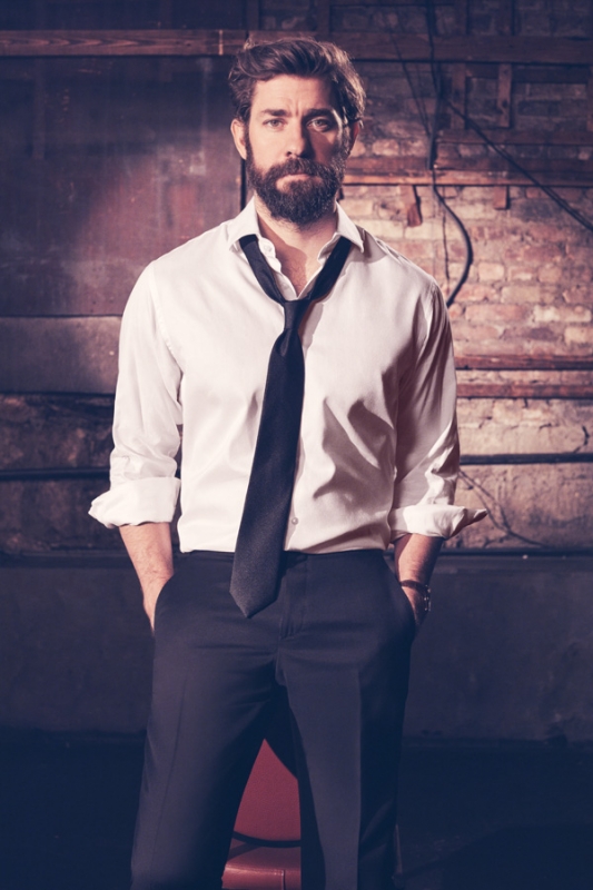 An adult male with a beard poses in a semi-formal outfit in front of a brick wall. He wears a white button-down shirt and black slacks.