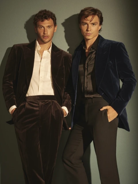 Two young adult men pose in velvet blazers and dress pants against a muted green background in a medium shot, showcasing formal fashion.