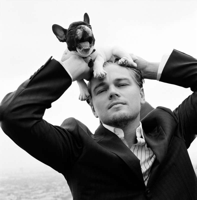 Black and white portrait of a man in a suit holding a French bulldog on his head outdoors with a blurred cityscape background.