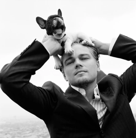 Black and white portrait of a man in a suit holding a French bulldog on his head outdoors with a blurred cityscape background.