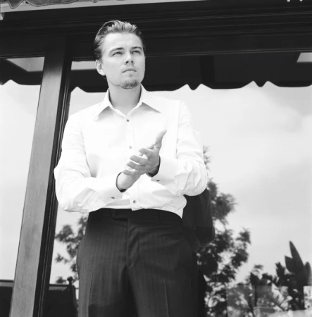 Leonardo DiCaprio in a white button-down shirt and dark trousers clapping outdoors, photographed by Nino Munoz.