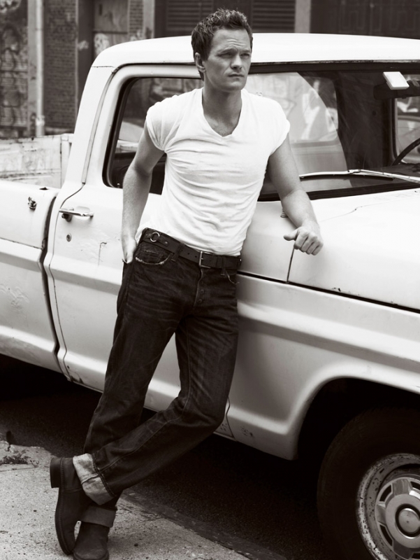 Black and white portrait of a man leaning against a vintage white pickup truck. The man wears a t-shirt, jeans, and boots.