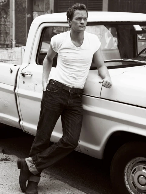Black and white portrait of a man leaning against a vintage white pickup truck. The man wears a t-shirt, jeans, and boots.