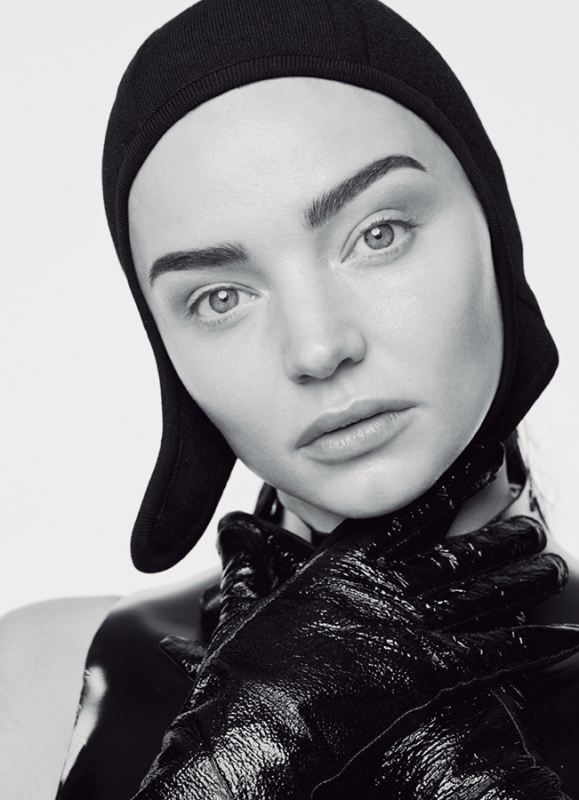 Black-and-white portrait of a woman with a black hat and shiny black gloves covering her neck, set against a clean, neutral backdrop.