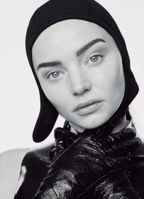 Black-and-white portrait of a woman with a black hat and shiny black gloves covering her neck, set against a clean, neutral backdrop.
