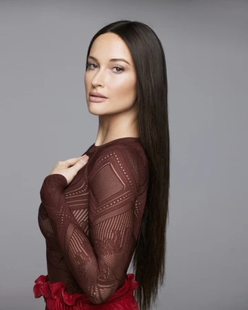Kacey Musgraves with long dark hair wearing a patterned burgundy top and red skirt against a grey background