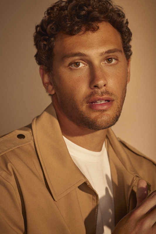 Portrait of an adult male with curly brown hair wearing a tan trench coat and white t-shirt against a neutral tan backdrop.
