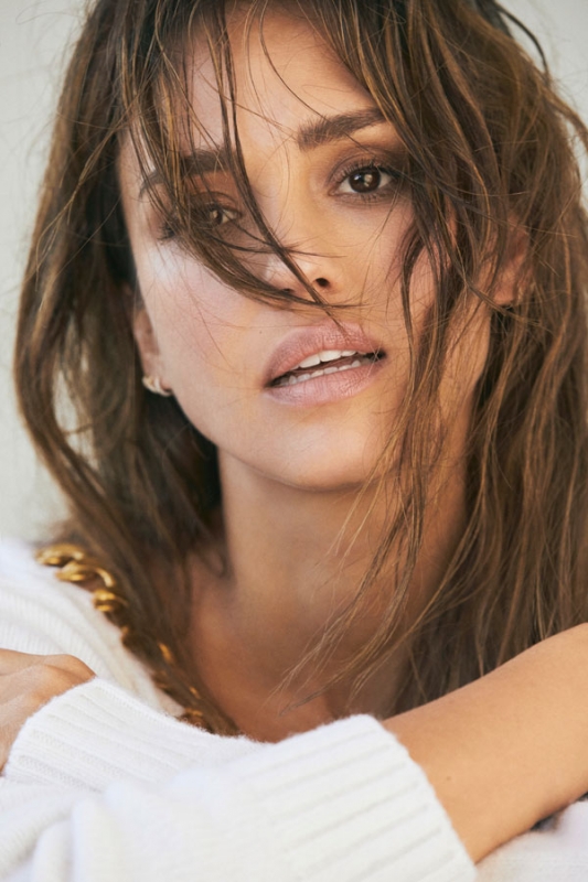 Close-up portrait of an adult female with light brown hair wearing a white sweater with a gold chain, gazing at the camera indoors.