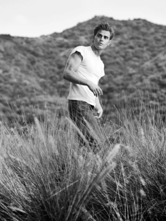 Black and white portrait of Paul Wesley standing in tall grass outdoors with a hilly landscape in the background