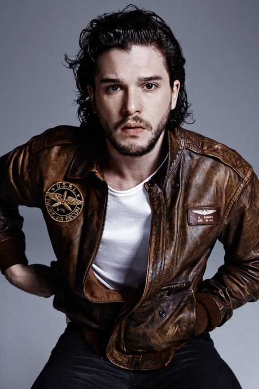 Portrait of a dark-haired male model wearing a brown leather aviator jacket and a white t-shirt against a grey studio backdrop.
