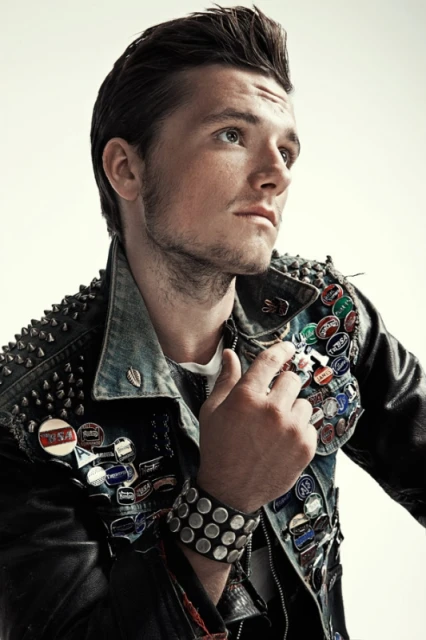 Portrait of a young adult male looking away, wearing a denim jacket with studs and pins, against a neutral light-grey backdrop.