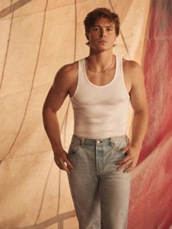 Young man in a white tank top and light wash denim jeans standing against a backdrop of draped fabric panels