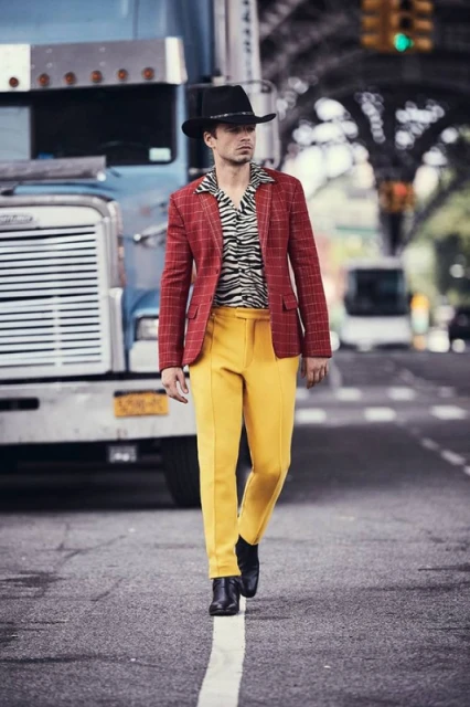 A man in a red plaid jacket and yellow pants walks on a city street, with a semi-truck in the background and city buildings.