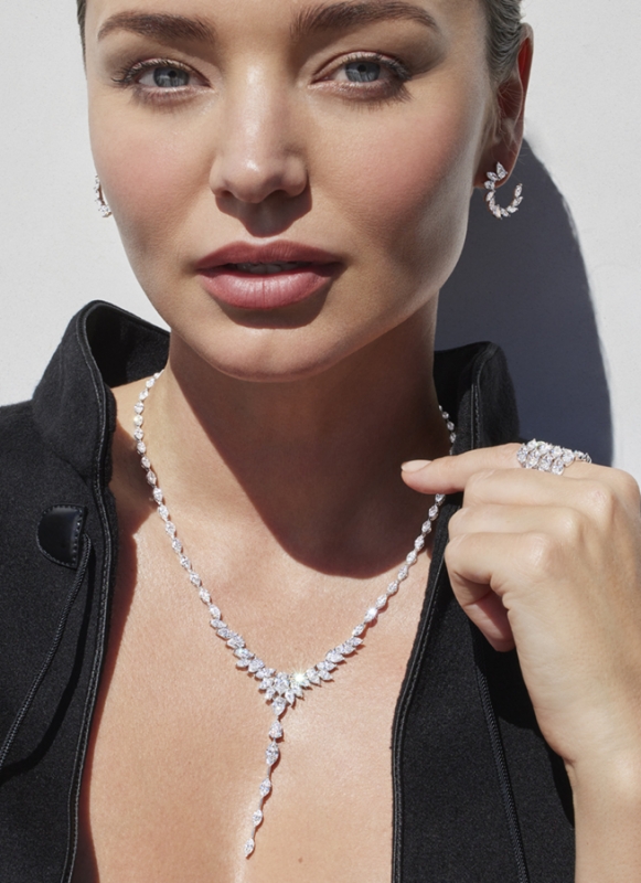 A woman models diamond jewelry, wearing earrings, a necklace, and a ring against a neutral background in a medium close-up shot.