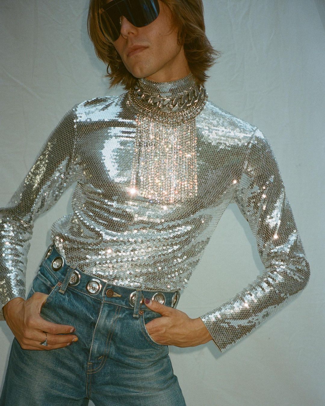 Man poses in a shiny silver long-sleeved top, light blue denim jeans with a silver grommet belt, dark glasses, and a heavy silver chain necklace.