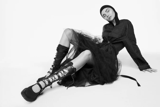 Model in black hooded top, tulle skirt, and lace-up boots sitting against a white background by Nino Munoz