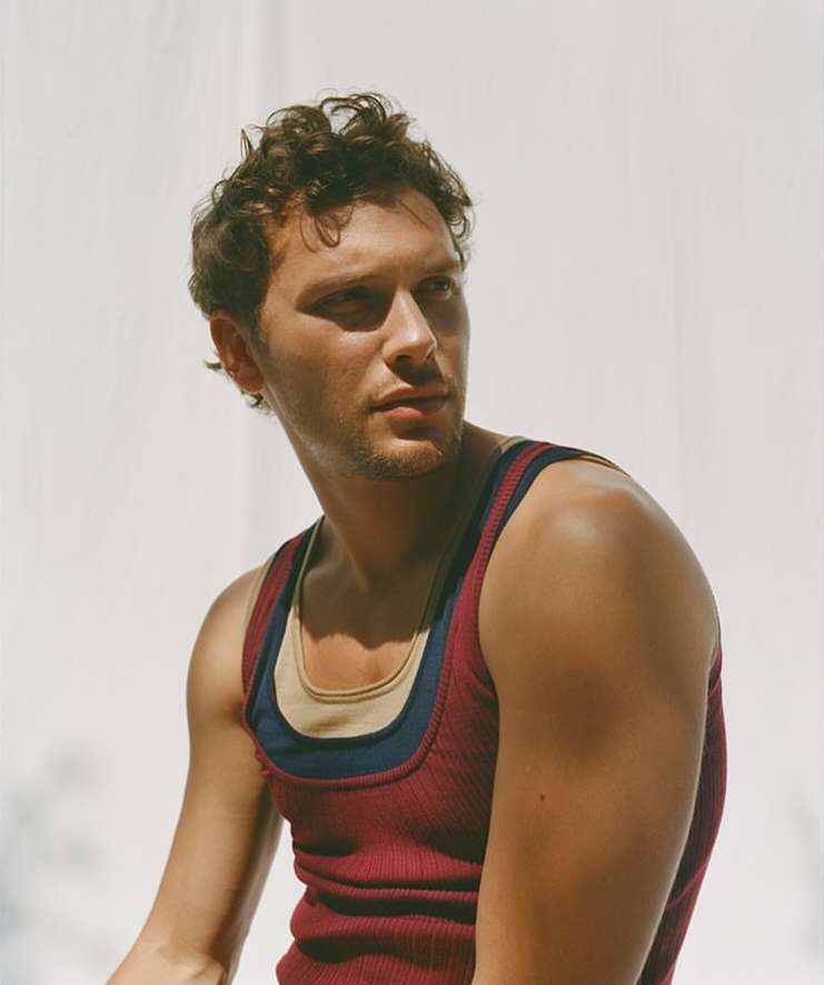 Portrait of a young man with curly brown hair, wearing a red tank top, posing in front of a white background with soft light and shadows.