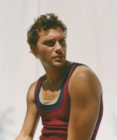 Portrait of a young man with curly brown hair, wearing a red tank top, posing in front of a white background with soft light and shadows.