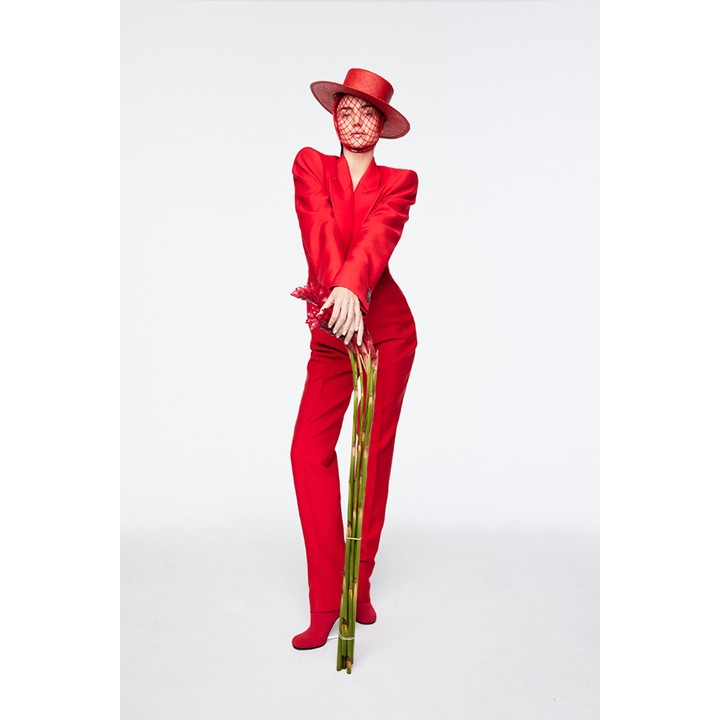 A stylish woman in a red suit and hat poses in a photo studio, holding a bouquet of bamboo stalks, against a bright white backdrop.