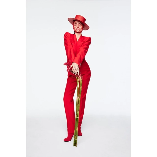A stylish woman in a red suit and hat poses in a photo studio, holding a bouquet of bamboo stalks, against a bright white backdrop.
