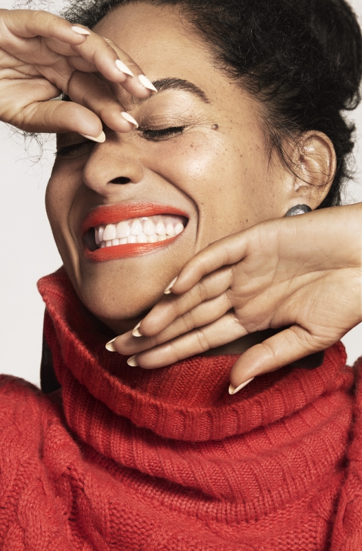 Close-up portrait of a smiling woman with curly hair and bright red lipstick, wearing a red turtleneck sweater against a white background.