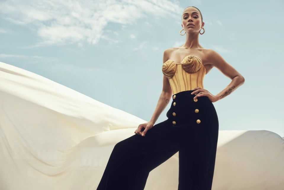 Zoe Saldana poses in a gold strapless top and black high-waisted pants against a bright sky and billowing white fabric backdrop.