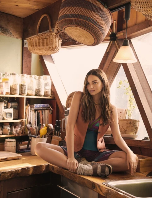 A woman with long brown hair sits cross-legged on a wooden countertop in a rustic kitchen. Baskets hang from the ceiling.