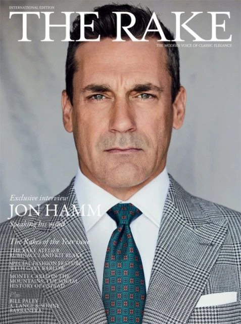 An adult male model poses in a photo studio wearing a grey suit with a blue shirt and a grey tie against a grey background.