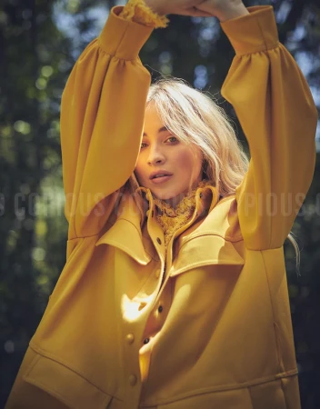 Blonde woman wearing a yellow jacket with her arms raised overhead outdoors