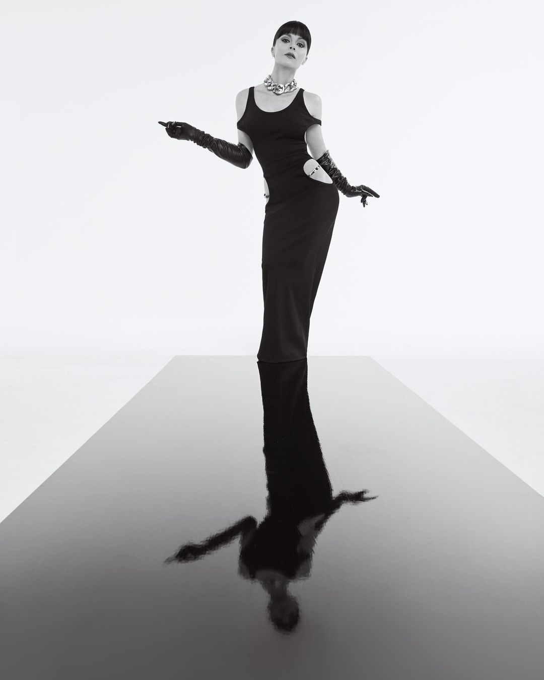 Elegant woman in a black gown and long gloves poses on a reflective black surface against a stark white background in a fashion photo.