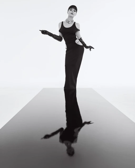Elegant woman in a black gown and long gloves poses on a reflective black surface against a stark white background in a fashion photo.