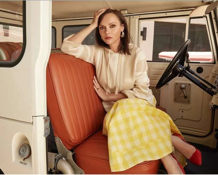 Christina Ricci sits in a vintage vehicle wearing a cream-colored hoodie and a yellow-and-white gingham skirt, posing for a fashion editorial.