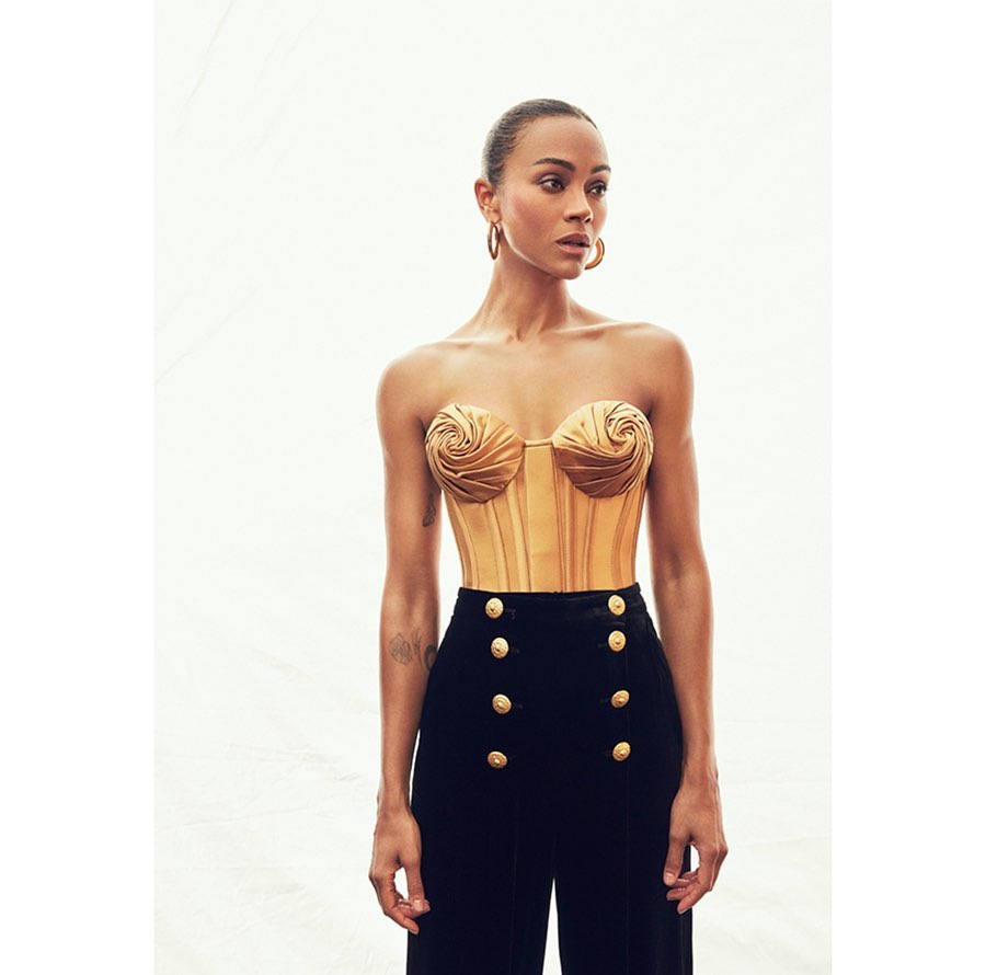 Zoe Saldana poses in a gold bustier and black high-waisted pants, against a light background in a studio-style setting, portrait shot.