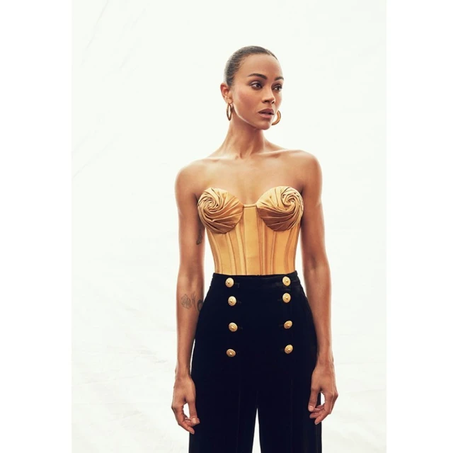 Zoe Saldana poses in a gold bustier and black high-waisted pants, against a light background in a studio-style setting, portrait shot.