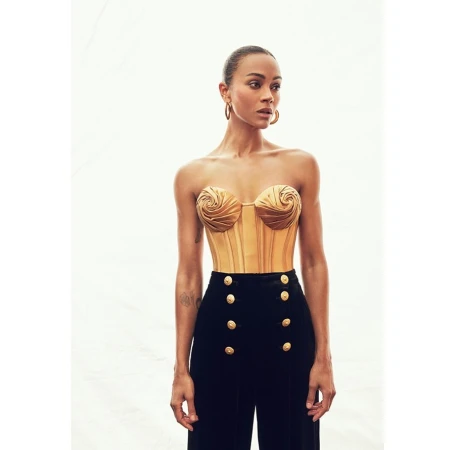 Zoe Saldana poses in a gold bustier and black high-waisted pants, against a light background in a studio-style setting, portrait shot.