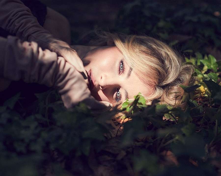 Portrait of a blonde woman lying down in a field of green plants in natural light. Her hands are near her face.