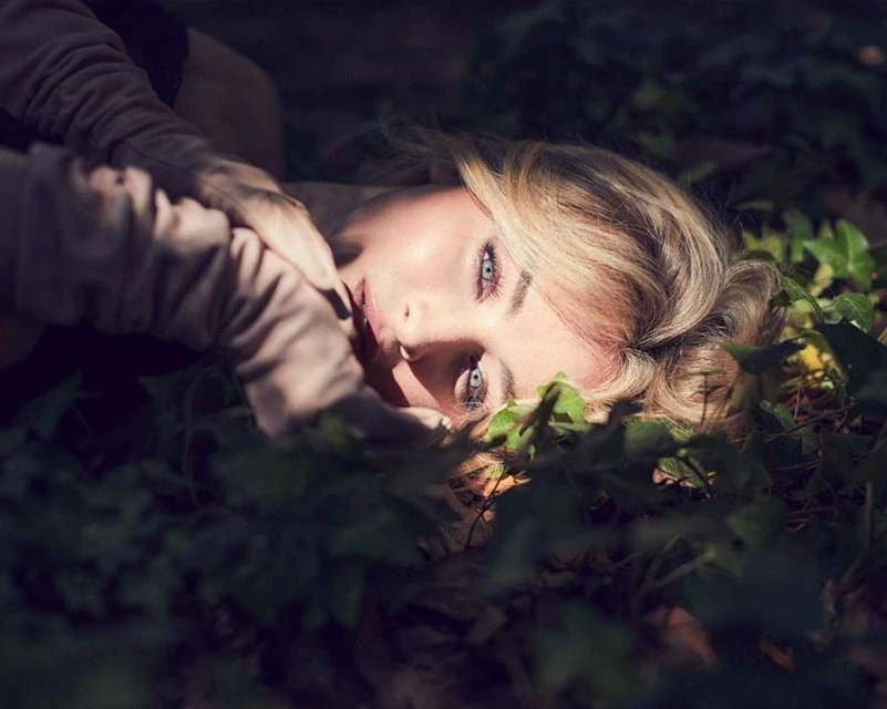 Portrait of a blonde woman lying down in a field of green plants in natural light. Her hands are near her face.