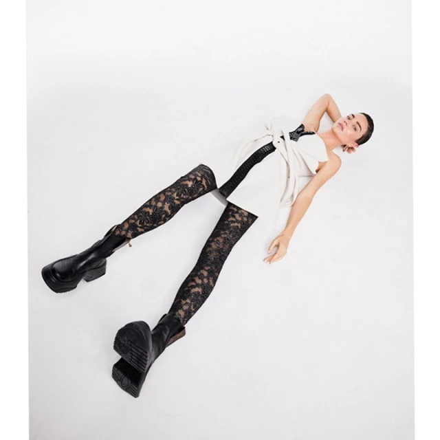 Woman in a mini dress, lace tights, and boots lies on a white surface in a studio setting, posing for a fashion shot.