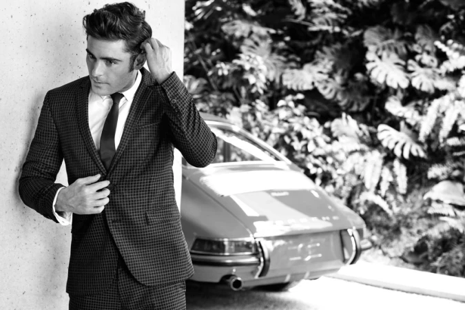 Zac Efron wearing a patterned suit leaning against a wall with a vintage car and foliage in the background