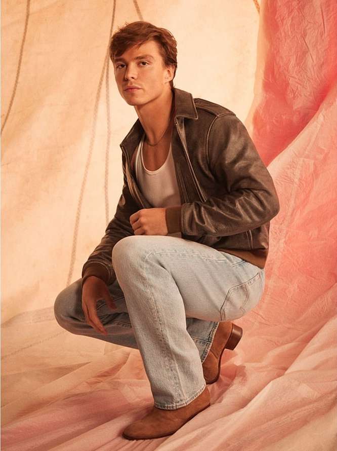 A young adult male, crouching in a studio, wearing a brown leather jacket, white t-shirt, and light blue jeans, posing against a pink backdrop.
