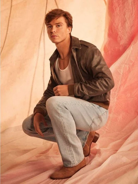 A young adult male, crouching in a studio, wearing a brown leather jacket, white t-shirt, and light blue jeans, posing against a pink backdrop.