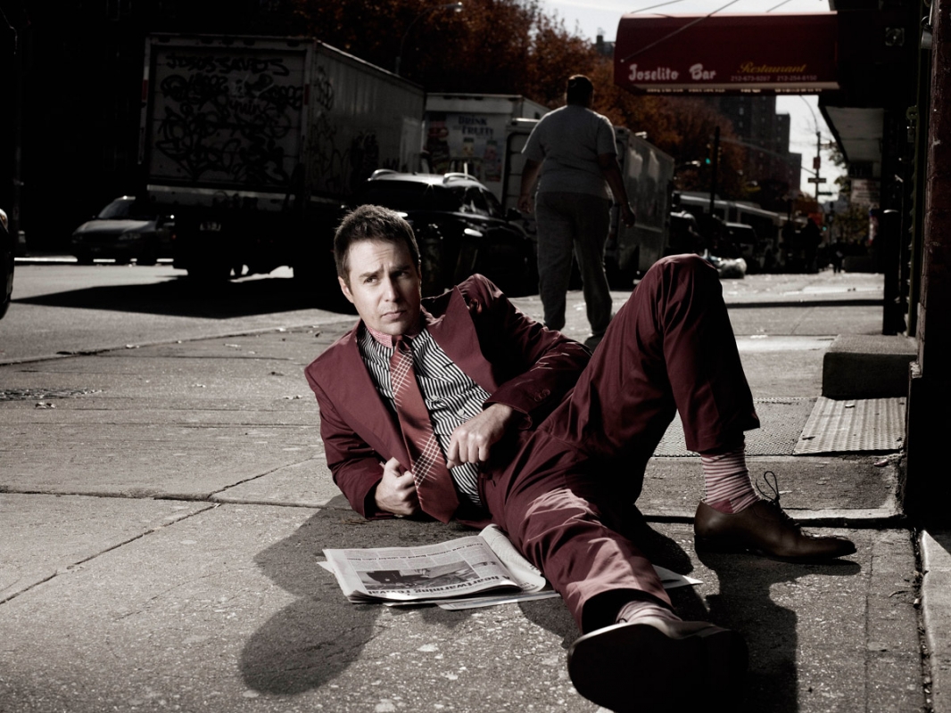 Man in a suit lying on a city sidewalk with a newspaper, with cars and buildings in the background, creating an editorial fashion image.